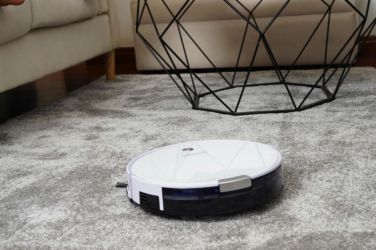 A white robot vacuum on a grey shaggy area rug under a wire-frame side table