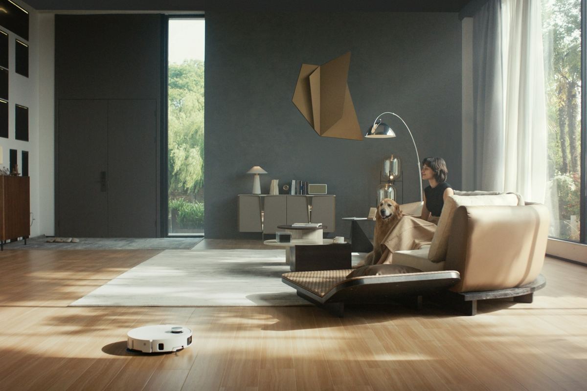 A small white robot vacuum mop on a wide wooden floor in a living room with large windows